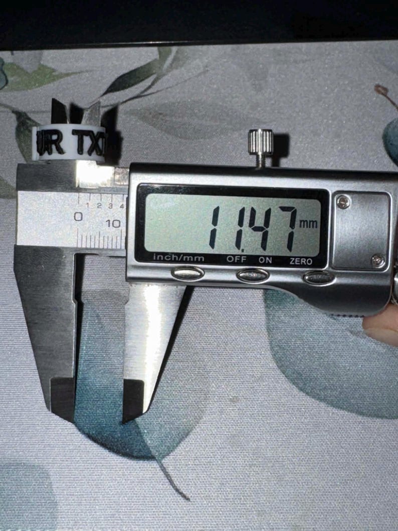 May include: A white plastic ring with the text "UR TX" is measured with a digital caliper. The caliper displays a measurement of 11.47 mm. The ring is positioned on a floral patterned surface.