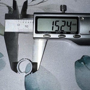 May include: A digital caliper measuring a small, light-colored ring. The measurement displayed is 15.24 mm, which is approximately 0.6 inches. The ring has a textured design around its outer edge. The background is a patterned fabric.