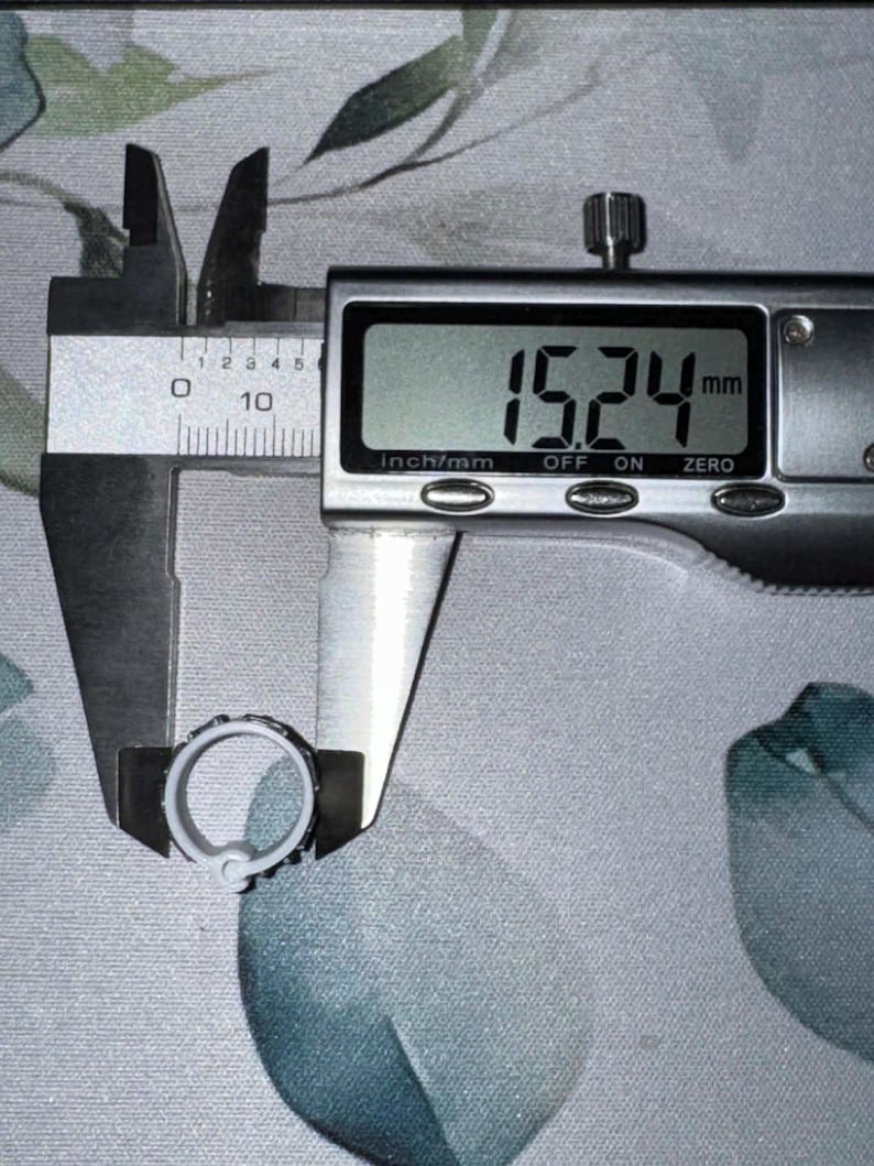 May include: A digital caliper measuring a small, light-colored ring. The measurement displayed is 15.24 mm, which is approximately 0.6 inches. The ring has a textured design around its outer edge. The background is a patterned fabric.