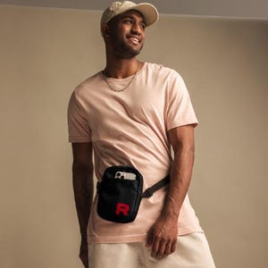 May include: A person wearing a light pink t-shirt with a black fanny pack with a red "R" on it. The fanny pack is worn around the waist and has a phone inside.