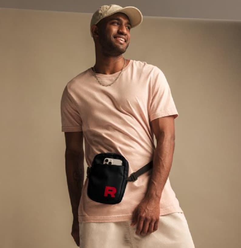 May include: A person wearing a light pink t-shirt with a black fanny pack with a red "R" on it. The fanny pack is worn around the waist and has a phone inside.