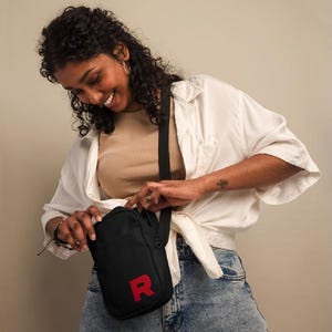 May include: A black crossbody bag with a red "R" on the front. The bag is being worn by a person with curly hair and a white shirt.