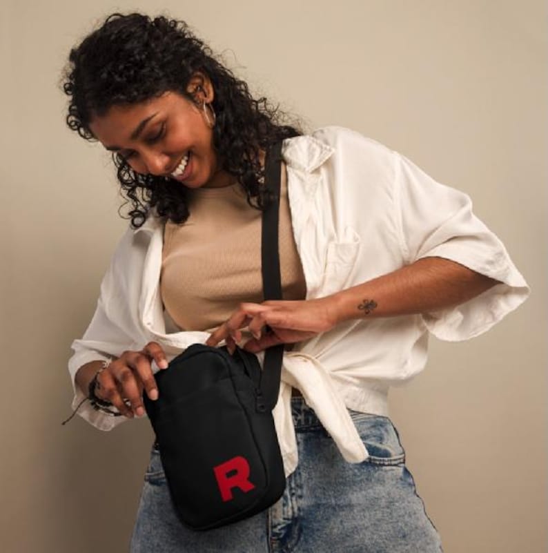 May include: A black crossbody bag with a red "R" on the front. The bag is being worn by a person with curly hair and a white shirt.