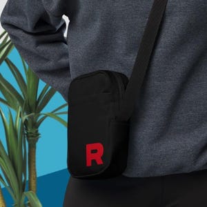 May include: Black crossbody bag with a red "R" on the front. The bag has a zipper closure and an adjustable strap.
