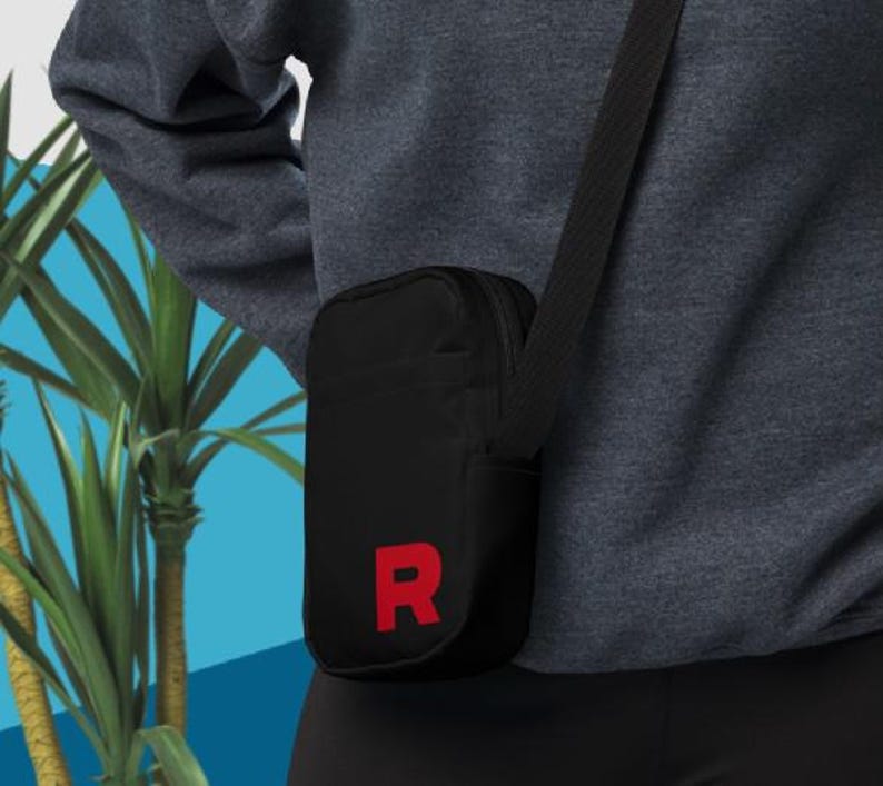 May include: Black crossbody bag with a red "R" on the front. The bag has a zipper closure and an adjustable strap.