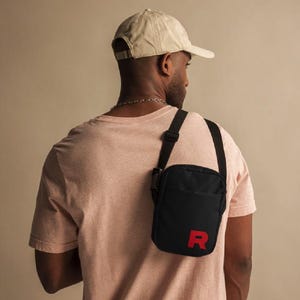 May include: A person wearing a black t-shirt and a beige baseball cap is carrying a black crossbody bag with a red "R" on the front.