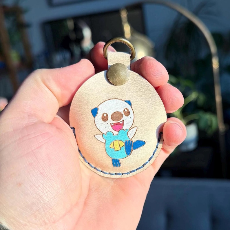 Puede incluir: Un llavero redondo de cuero beige con un personaje de nutria de dibujos animados con detalles en azul y amarillo. El llavero tiene un anillo y un cierre de bot&oacute;n color lat&oacute;n, con costuras azules alrededor del borde.
