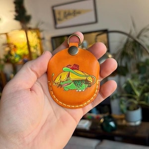 May include: Orange leather keychain featuring a hand-painted illustration of a green and yellow cartoon character. The keychain has a metal ring and a stitched border. The design is a popular character from a video game.