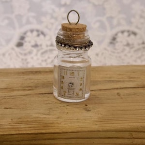 Puede incluir: Pequeña botella de vidrio transparente con tapón de corcho y anillo decorativo de metal. La botella presenta una etiqueta con un diseño de esfera de reloj vintage y una pequeña joya. Se muestra sobre una superficie de madera.