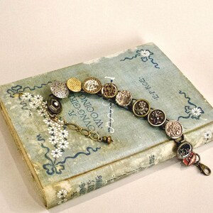 Puede incluir: Un brazalete vintage con varios botones metálicos y decorativos. El brazalete se muestra sobre un libro azul claro con detalles florales y de texto. Tiene una cadena y un cierre dorados.