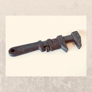 May include: A vintage adjustable wrench, crafted from dark, aged metal. The tool features a long handle with a circular hole at the end and a movable jaw for adjusting to different sizes. The wrench is designed for tightening or loosening nuts and bolts.