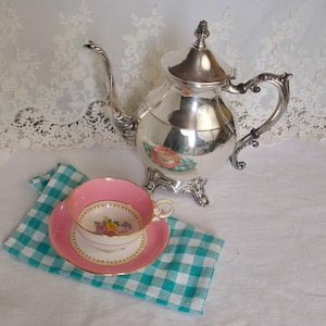 Puede incluir: Un juego de té plateado con una tetera, una taza y un platillo. La taza y el platillo son rosas con un diseño floral. El juego está sobre una servilleta a cuadros azul y blanco.