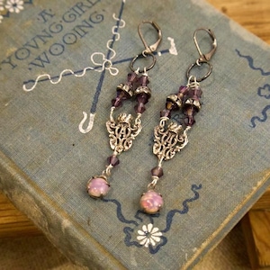 Puede incluir: Un par de pendientes colgantes con detalles en metal plateado y cuentas moradas. Los pendientes presentan diseños de filigrana ornamentados y piedras rosas en forma de lágrima. Se encuentran sobre un libro antiguo.