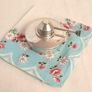 Puede incluir: Una lámpara de aceite de metal plateado con asa descansa sobre un pañuelo floral azul claro. El pañuelo tiene un borde festoneado y presenta flores rosas y blancas. El fondo es una superficie blanca.