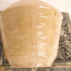 Puede incluir: Un cuenco de cerámica de color beige claro con un acabado brillante y un patrón de tejido de cesta texturizado en relieve en la superficie exterior. El cuenco tiene una forma redondeada con una base ligeramente cónica y un color uniforme.