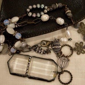 Può includere: Una collana con un ciondolo in vetro rettangolare trasparente con una fila centrale di strass, collegata a una catena di perle bianche, perline iridescenti, pietre grigie, maglie in metallo color bronzo, ciondoli a nodo e una medaglia circolare.