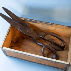 May include: A pair of large, weathered metal shears with a dark, oxidized finish resting inside a rectangular wooden box. The box features light-colored wood panels with dark, decorative trim along the edges and a visible grain pattern.
