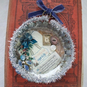 Könnte beinhalten: Ein kreisförmiges Ornament aus einer Metall-Tarteform mit silbernem Lamellarand und einer blauen Samtschleife. Das Innere enthält eine Vintage-Weihnachtskarte mit dem Text Christmas Joys to You, ein kleines Zifferblatt, Knöpfe und blaue Cabochons.