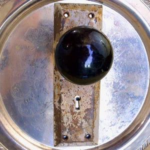 Puede incluir: Un pomo de puerta negro vintage y una placa de cerradura están montados en una bandeja plateada. La bandeja tiene un diseño ornamental en relieve y un borde decorativo en forma de cuerda. La ferretería de la puerta muestra signos de edad y desgaste.
