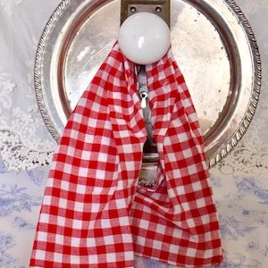 May include: A vintage-style silver serving tray with a decorative rim, a white ceramic knob, and a red and white checkered kitchen towel hanging from it. The towel is folded and draped over the knob.