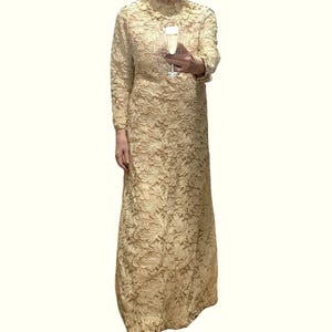 May include: A floor-length, long-sleeved dress in a pale gold hue, featuring an intricate, raised floral lace pattern throughout the entire garment. The dress has a high neckline and a straight, column-like silhouette that extends to the floor.