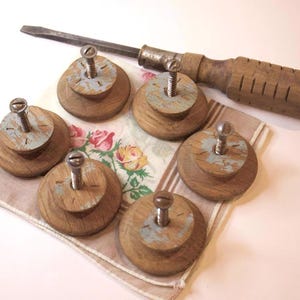Puede incluir: Seis pomos de madera vintage con tornillos metálicos, dispuestos sobre un pañuelo con estampado floral. Un destornillador vintage con mango de madera también está presente. Los pomos tienen un aspecto envejecido y desgastado.