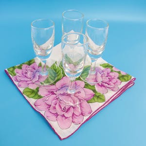 Könnte beinhalten: Vier klare Schnapsgläser auf einem weißen, floralen Taschentuch mit rosa und lila Blumen und grünen Blättern. Das Taschentuch hat einen rosa Rand. Der Hintergrund ist einfarbig hellblau.