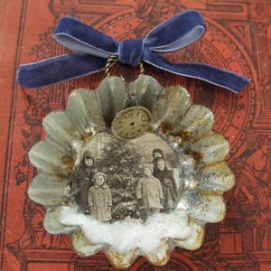 May include: A decorative ornament featuring a fluted metal tart mold with a rusted finish, containing a black and white photograph of four children near a tree. A small clock face charm and a blue velvet ribbon bow are attached to the top.