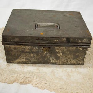 Puede incluir: Una caja de seguridad rectangular de metal con un acabado gris oscuro desgastado y signos visibles de óxido. La caja cuenta con una tapa abatible con un asa central de metal y un mecanismo de cerradura en el panel frontal.