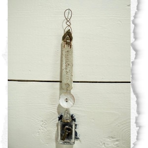 May include: A vertical hanging ornament featuring a vintage metal bottle opener stamped with the text REGAL.NER, attached to a round white mother-of-pearl button and a small clear glass bottle containing a dark metal key and blue fabric.