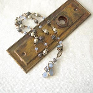 Könnte beinhalten: Eine Halskette mit einer Kette aus Metallperlen, filigranen Kugeln und kleinen hellblauen Kristallen. Die Kette endet in einem Anhänger aus größeren, glatten, hellblauen tropfenförmigen Steinen, die auf einer rechteckigen Messingplatte liegen.