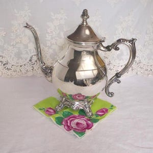 May include: A polished silver teapot featuring a curved spout, a decorative handle, and a lid with a finial top. The teapot stands on three ornate feet and has a reflective surface that shows the surrounding environment and light.
