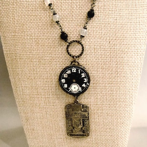 Può includere: Una collana caratterizzata da una catena con perline alternate nere e bianche traslucide, un anello in metallo intrecciato, un quadrante circolare nero con numeri bianchi e un ciondolo rettangolare in metallo color bronzo con testo in rilievo.
