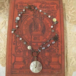 May include: A necklace featuring a large, round, white mother-of-pearl button pendant suspended from a bronze-toned metal chain. The chain is accented with small white pearls, translucent grey beads, and a decorative metal floral-shaped link.
