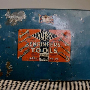 Puede incluir: Caja de herramientas de metal azul vintage con signos de desgaste. La caja de herramientas tiene una etiqueta naranja con el texto "NURO ENGINEERS TOOLS" e ilustraciones de herramientas. Una pequeña bandera es visible.
