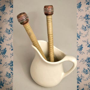 Könnte beinhalten: Zwei antike Holzbobbins mit roten und silbernen Akzenten, die in einem weißen Keramikkrug stehen. Die Spulen haben eine zylindrische Form mit einem strukturierten Design. Der Krug hat einen gebogenen Griff und eine glatte Oberfläche.