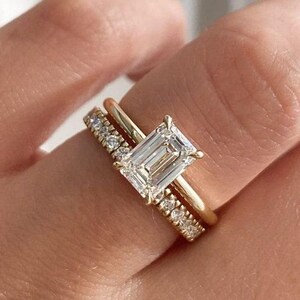 Op de afbeelding: Twee gouden ringen worden op een vinger getoond. De hoofdring heeft een grote, rechthoekige, heldere edelsteen. De tweede ring is een gouden band met kleine, heldere edelstenen. De ringen staan tegen een zachte, neutrale achtergrond.