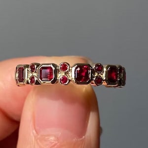 Può includere: Un anello d'oro con una serie di pietre preziose rosse quadrate e rotonde alternate. Le pietre sono incastonate in un motivo ripetuto attorno alla fascia. Lo sfondo è grigio.