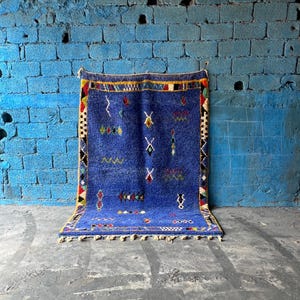 Op de afbeelding: Een blauw Marokkaans tapijt met een geometrisch patroon in rood, geel en wit. Het tapijt heeft een franje rand en hangt aan een blauwe bakstenen muur.