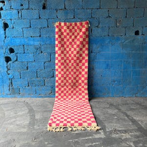 May include: A pink and cream checkered rug with fringe. The rug is hanging on a blue brick wall.