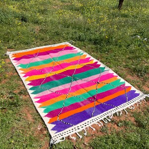 Pode incluir: Um tapete retangular e colorido com um padr&atilde;o geom&eacute;trico. O tapete apresenta listras horizontais em tons de rosa, laranja, verde e roxo, com uma borda branca e borlas. Formas de diamante em preto e branco s&atilde;o tecidas no design.