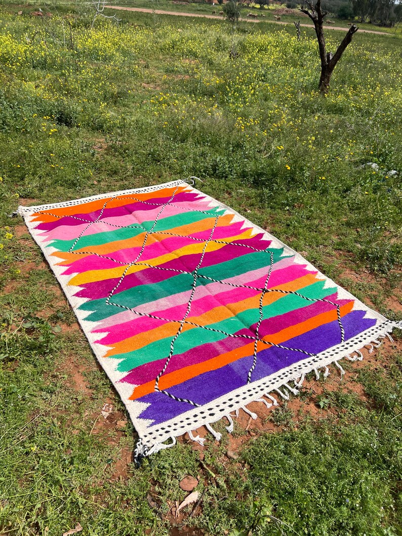 Pode incluir: Um tapete retangular e colorido com um padr&atilde;o geom&eacute;trico. O tapete apresenta listras horizontais em tons de rosa, laranja, verde e roxo, com uma borda branca e borlas. Formas de diamante em preto e branco s&atilde;o tecidas no design.
