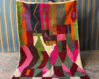 May include: A colorful Moroccan rug with geometric patterns in shades of pink, green, red, orange, and purple. The rug has a white background and is made of wool.