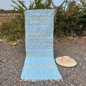 May include: A rectangular, light blue rug with a repeating pattern of yellow geometric shapes. The rug has a white fringe on the bottom edge and a red border. A straw hat rests on the ground nearby.