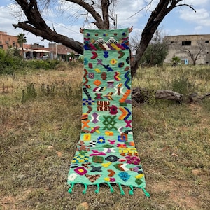 May include: A long, rectangular, hand-woven rug with a light green background and colorful geometric patterns. The rug features various shapes in shades of orange, blue, yellow, and pink. Green tassels adorn the bottom edge. The rug is displayed outdoors.