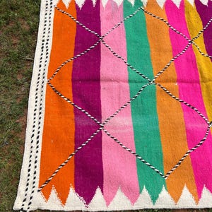 Pode incluir: Um tapete vibrante, tecido &agrave; m&atilde;o, com um design listrado colorido. O tapete apresenta listras verticais em tons de laranja, f&uacute;csia, rosa, verde e amarelo, intersectadas por um padr&atilde;o de losangos. As bordas s&atilde;o finalizadas com uma borda branca e borlas.