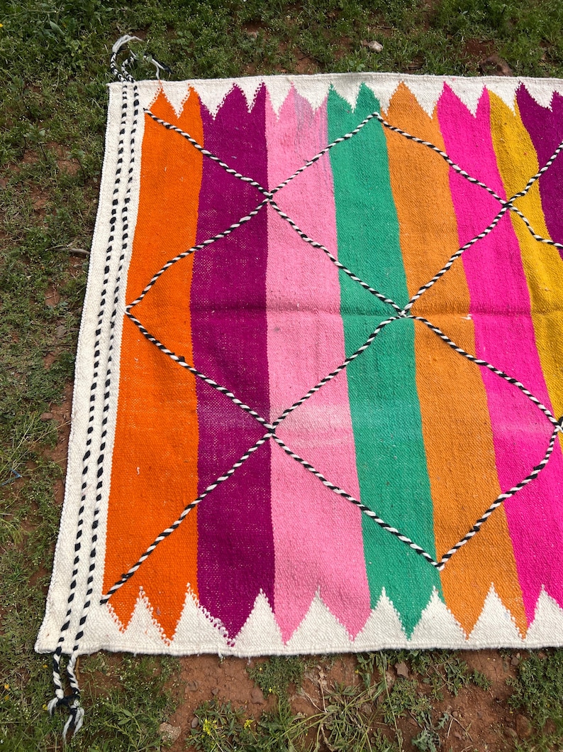 Pode incluir: Um tapete vibrante, tecido &agrave; m&atilde;o, com um design listrado colorido. O tapete apresenta listras verticais em tons de laranja, f&uacute;csia, rosa, verde e amarelo, intersectadas por um padr&atilde;o de losangos. As bordas s&atilde;o finalizadas com uma borda branca e borlas.