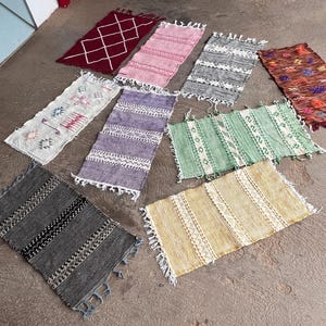 Może przedstawiać: Kolekcja małych, prostokątnych dywaników w różnych kolorach i wzorach. Dywaniki mają wzory w odcieniach czerwieni, różu, szarości, bieli, fioletu, zieleni i żółci. Każdy dywanik ma frędzel na krawędzi, a wzory są różne, od geometrycznych po paski.