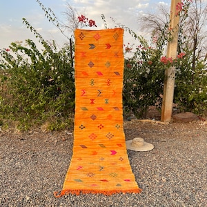 Op de afbeelding: Een oranje loper met geometrische patronen in verschillende kleuren. De loper is rechthoekig met een gefranjerde rand. Het ontwerp omvat pijlen, ruiten en andere vormen. De loper wordt buiten tentoongesteld.