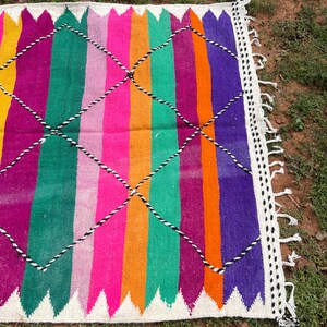 Pode incluir: Um tapete retangular vibrante com listras verticais em amarelo, rosa, laranja, verde, roxo e magenta. O tapete apresenta uma borda branca com um padr&atilde;o em zigue-zague e detalhes em diamante preto e branco.
